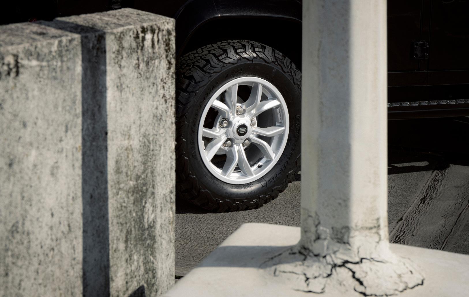Close-up of a tire and silver wheel near cracked concrete pillar.