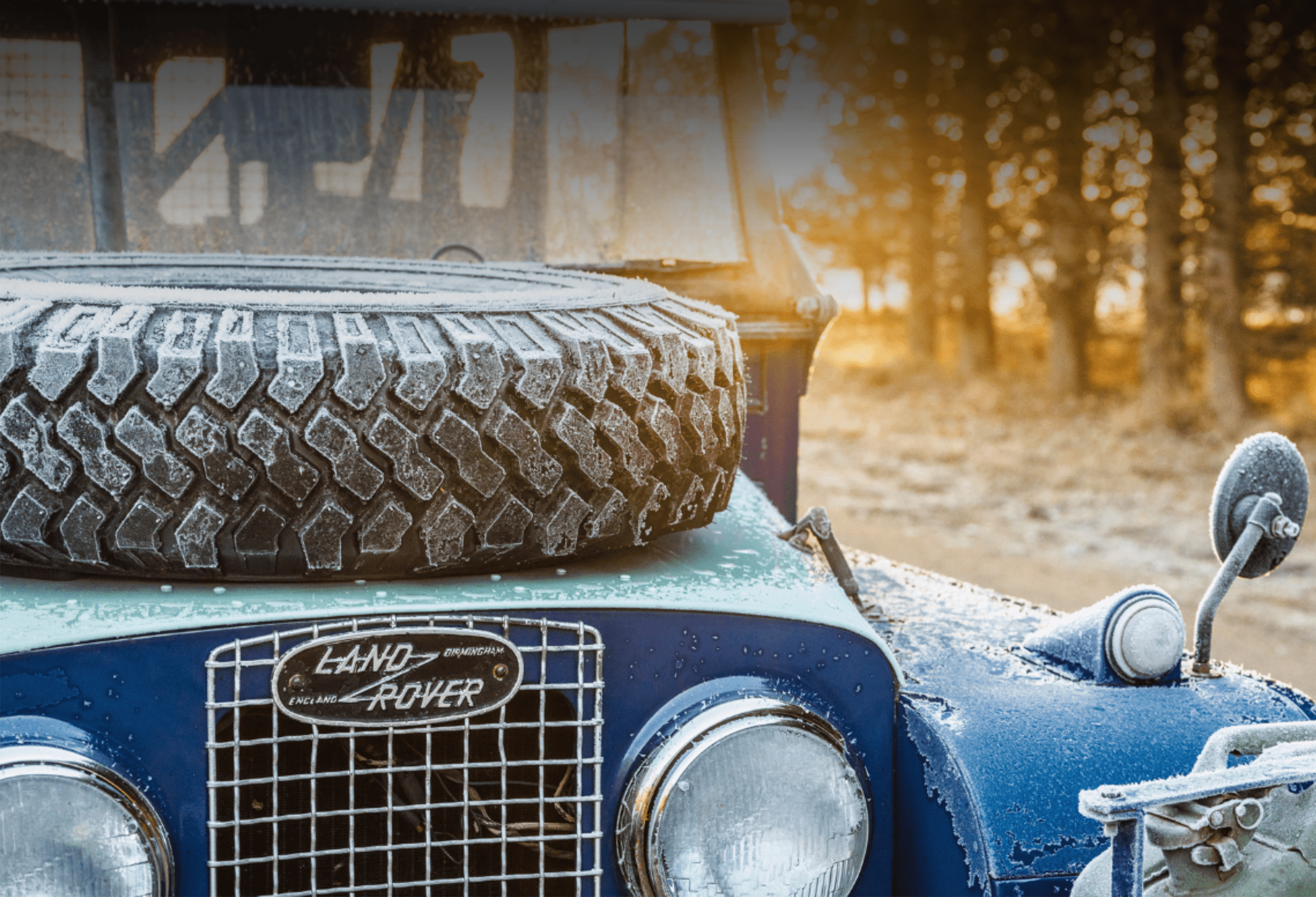 Frost-covered vintage Land Rover with hood-mounted spare tire in forest setting.