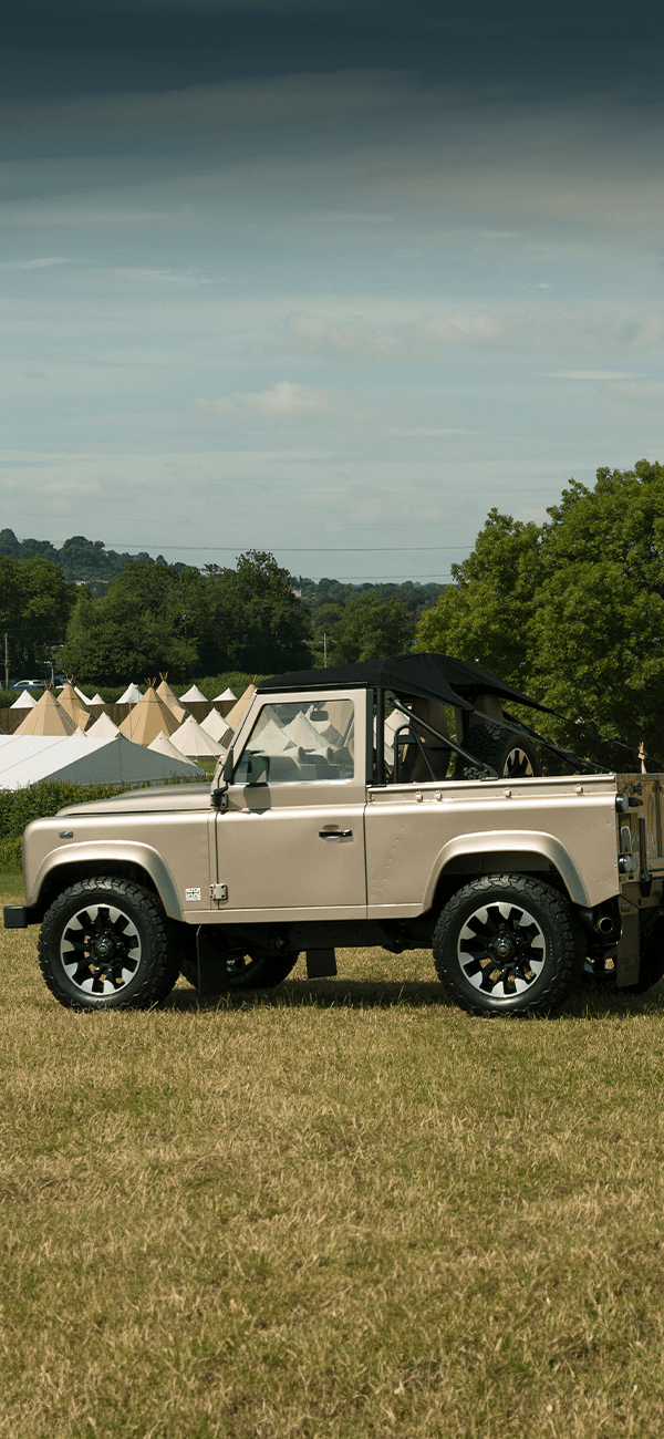 Two off-road Defenders parked on a grassy field with tents and trees in the background.