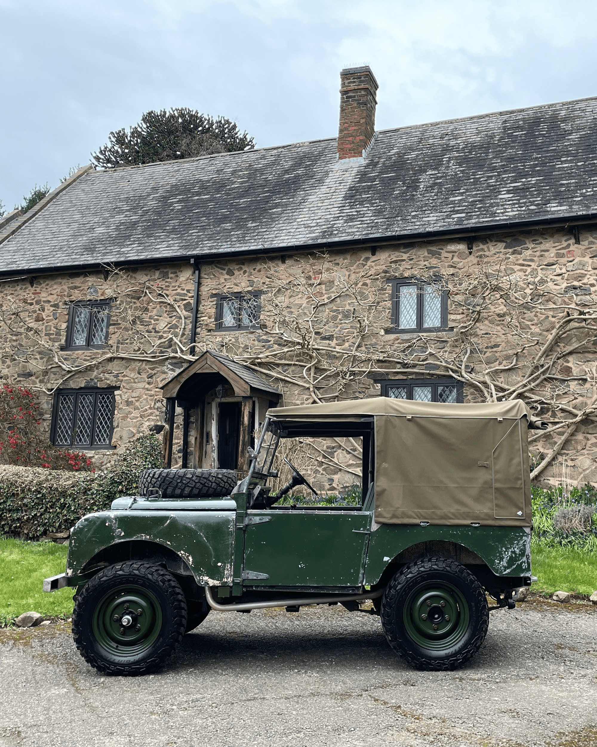 Classic green off-road Land Rover with soft top parked in front of stone house with climbing plants.