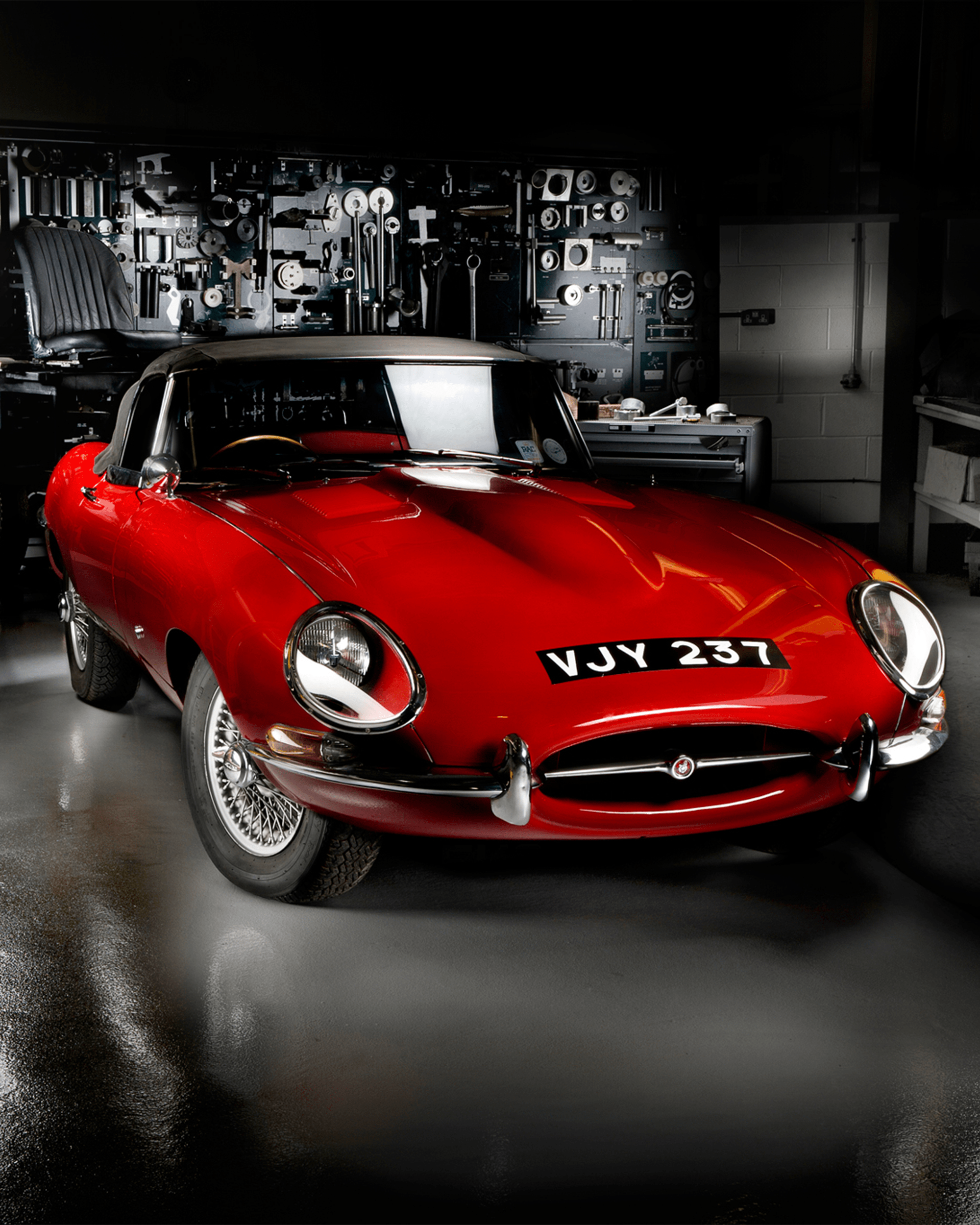 Classic red Jaguar E-Type parked in a workshop with tools on the wall.