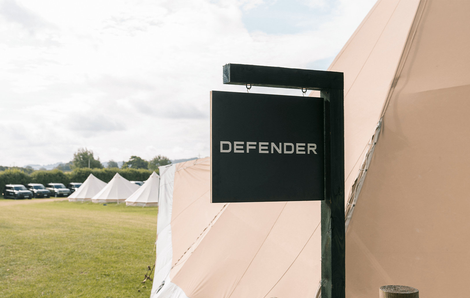 DEFENDER sign at a grassy campsite with tents and Defenders in the background.
