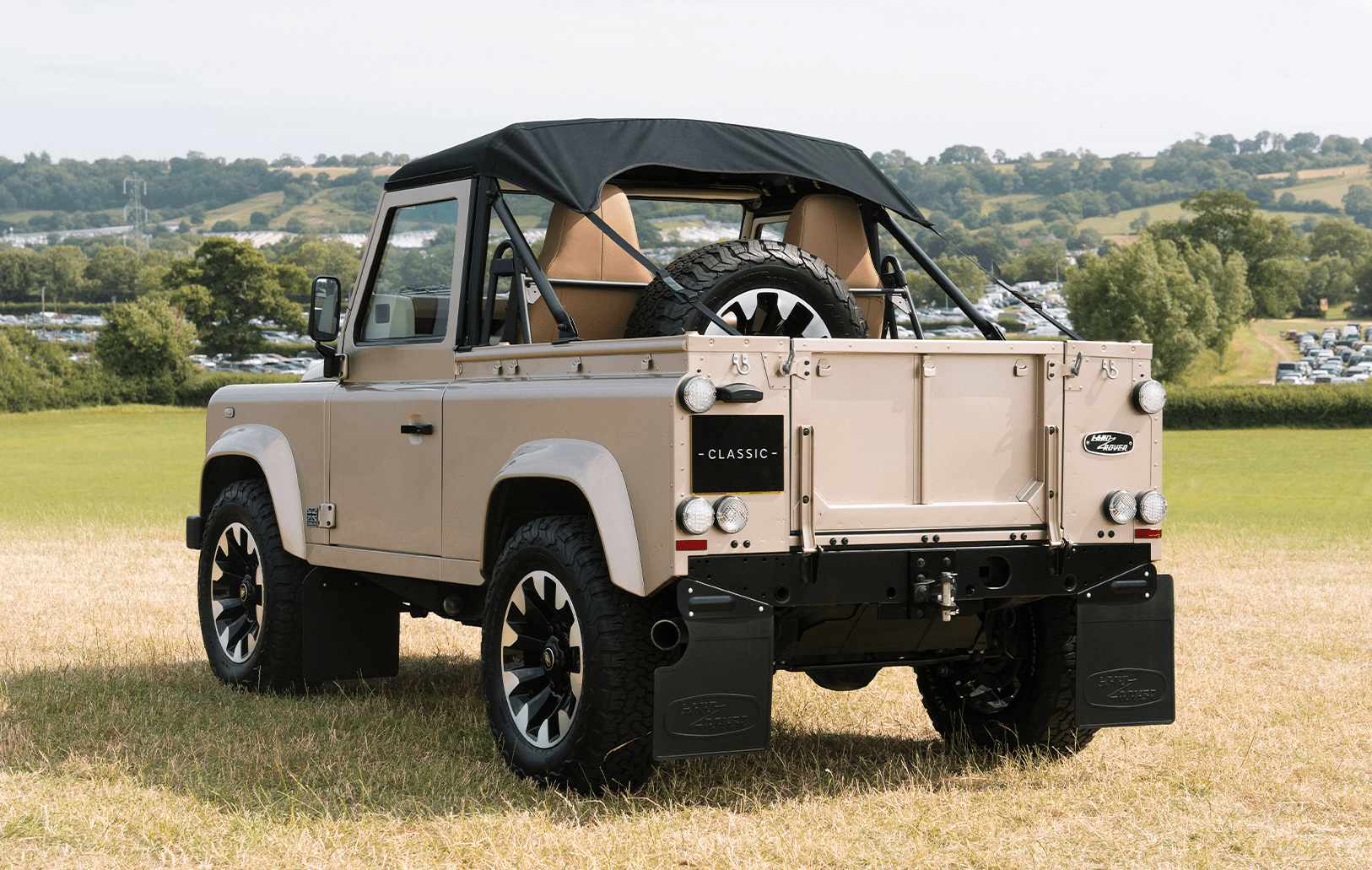 Beige Land Rover Defender with soft top and spare tire, viewed from rear three-quartre angle on grass.