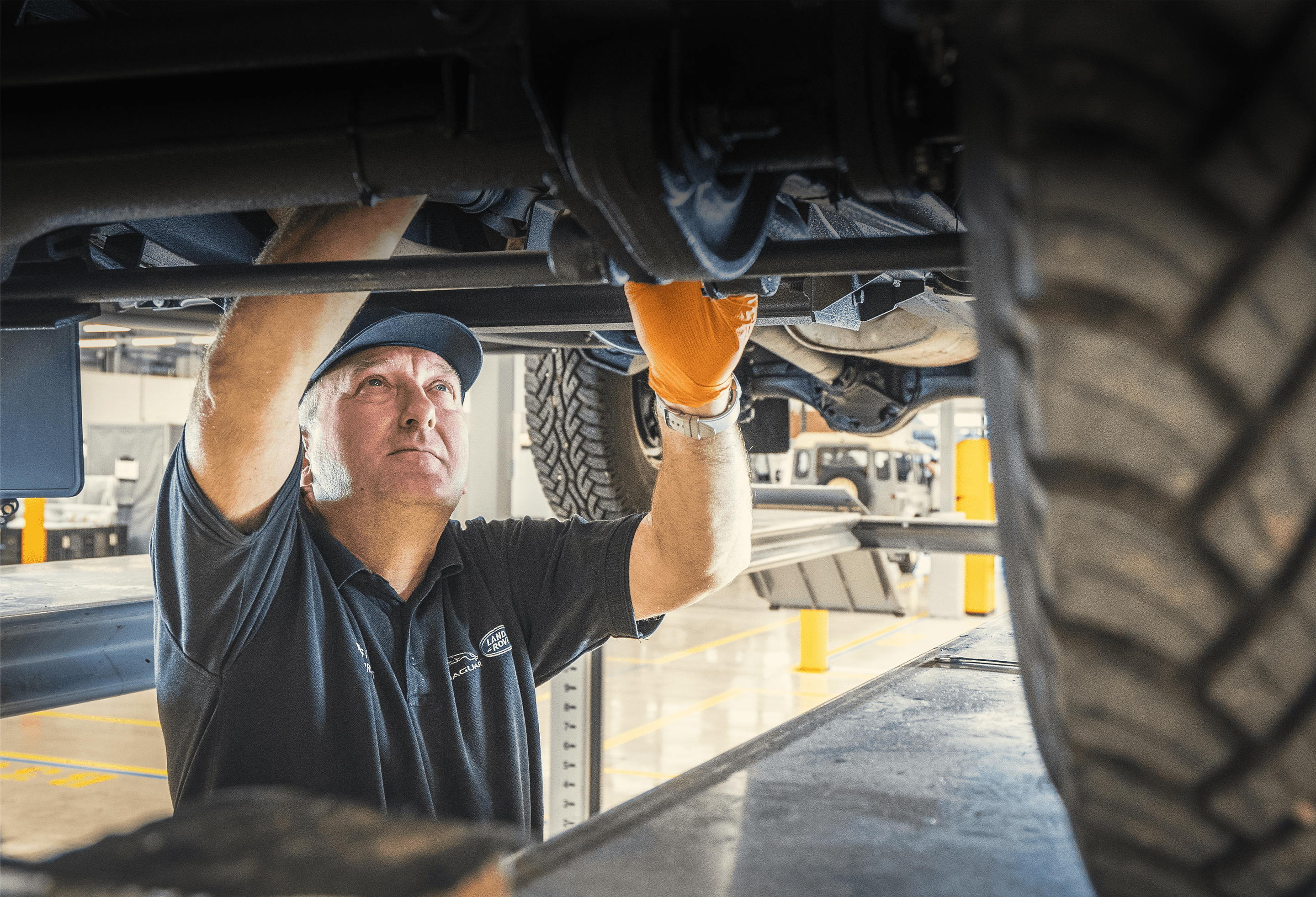 Person in orange gloves working under a vehicle in an automotive workshop.
