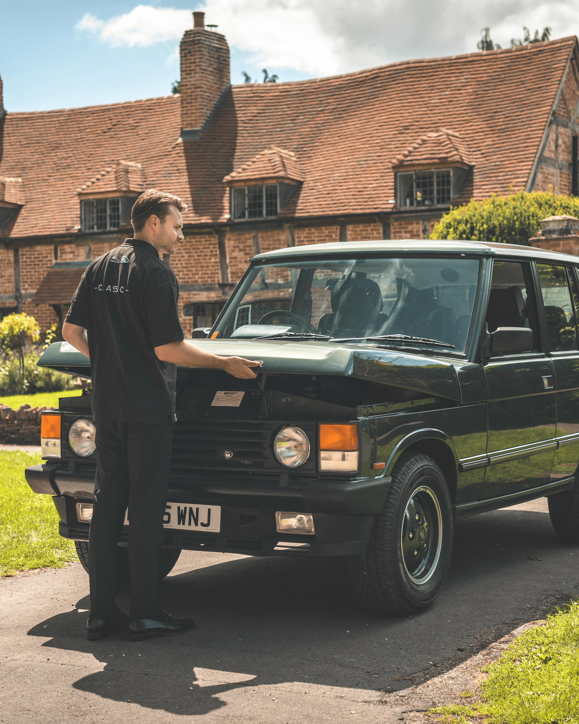 Mechanic inspecting Range Rover with open hood outside large brick house.