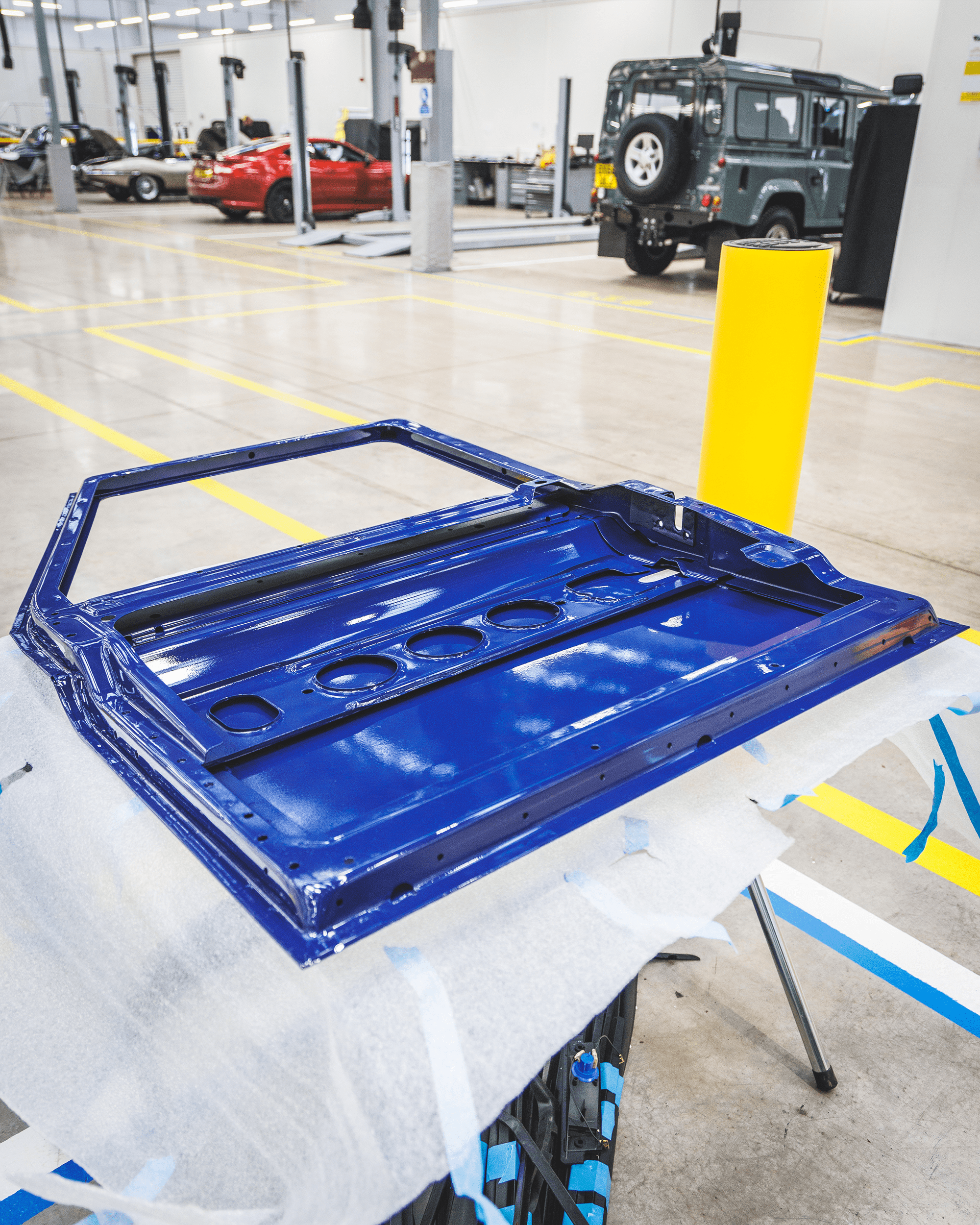 Blue car door panel on workbench in workshop with vehicles and tools in background.