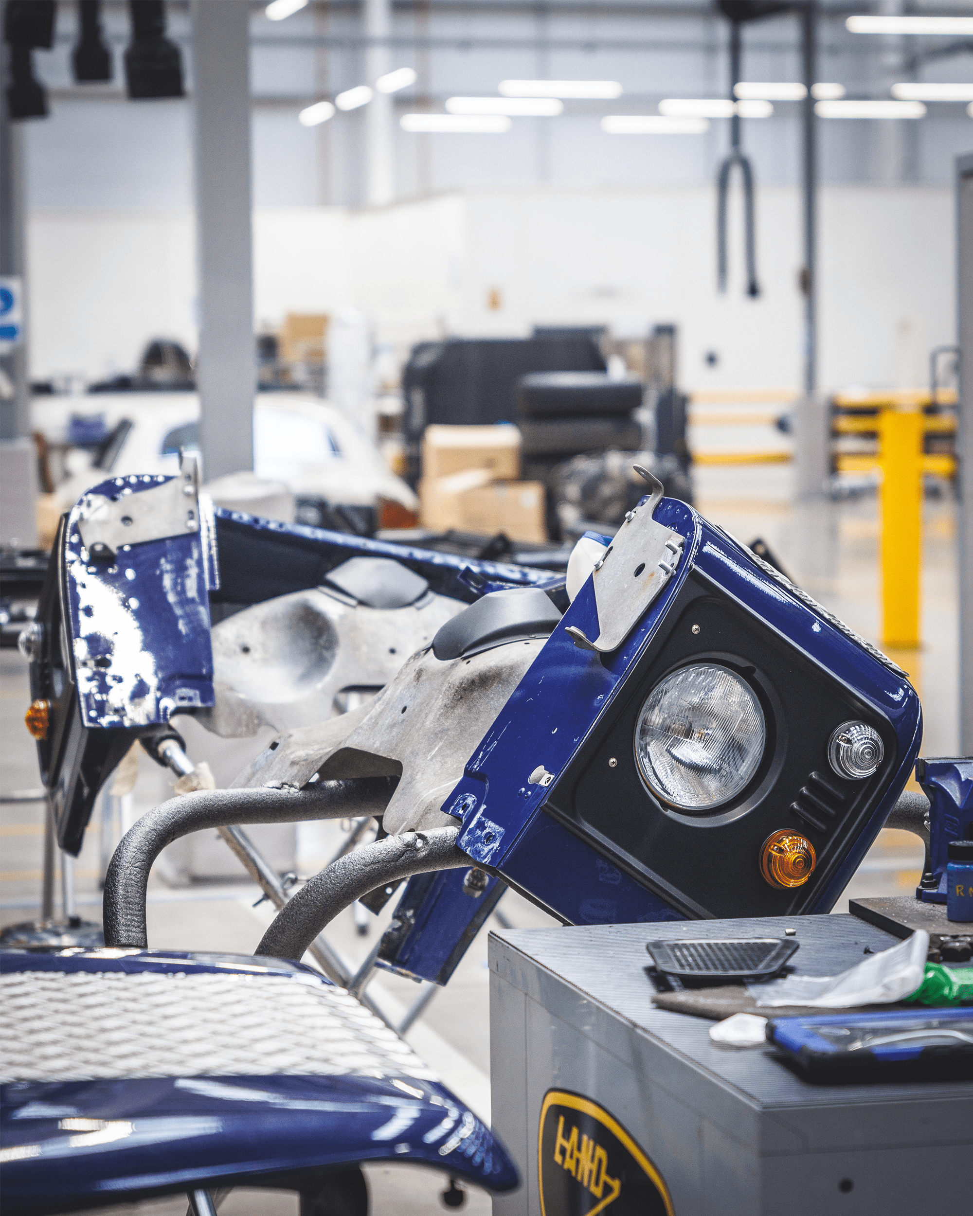 Blue vehicle front panel with headlight in a workshop filled with car parts and tools.
