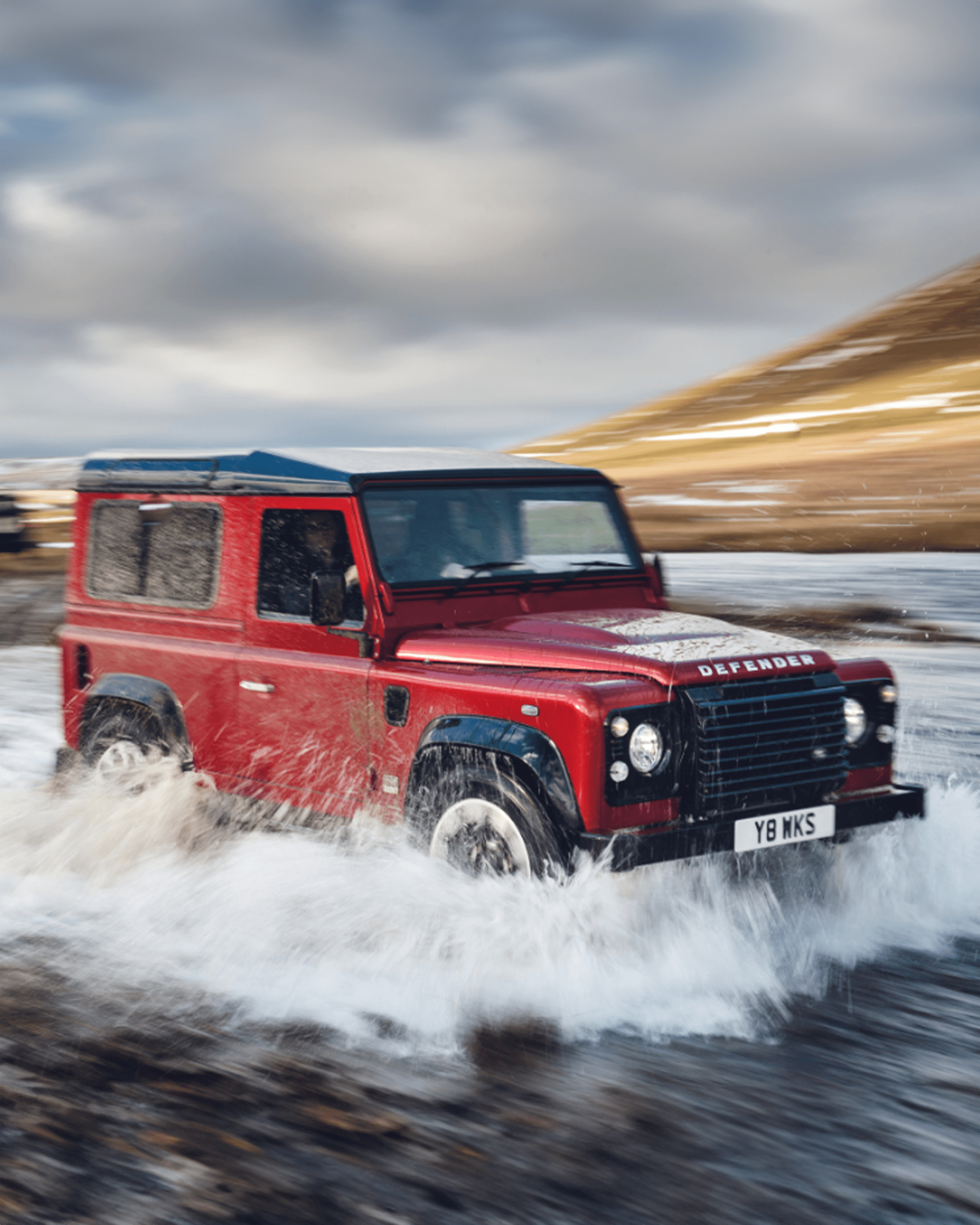 Red Land Rover Defender driving through water with splashes around wheels.