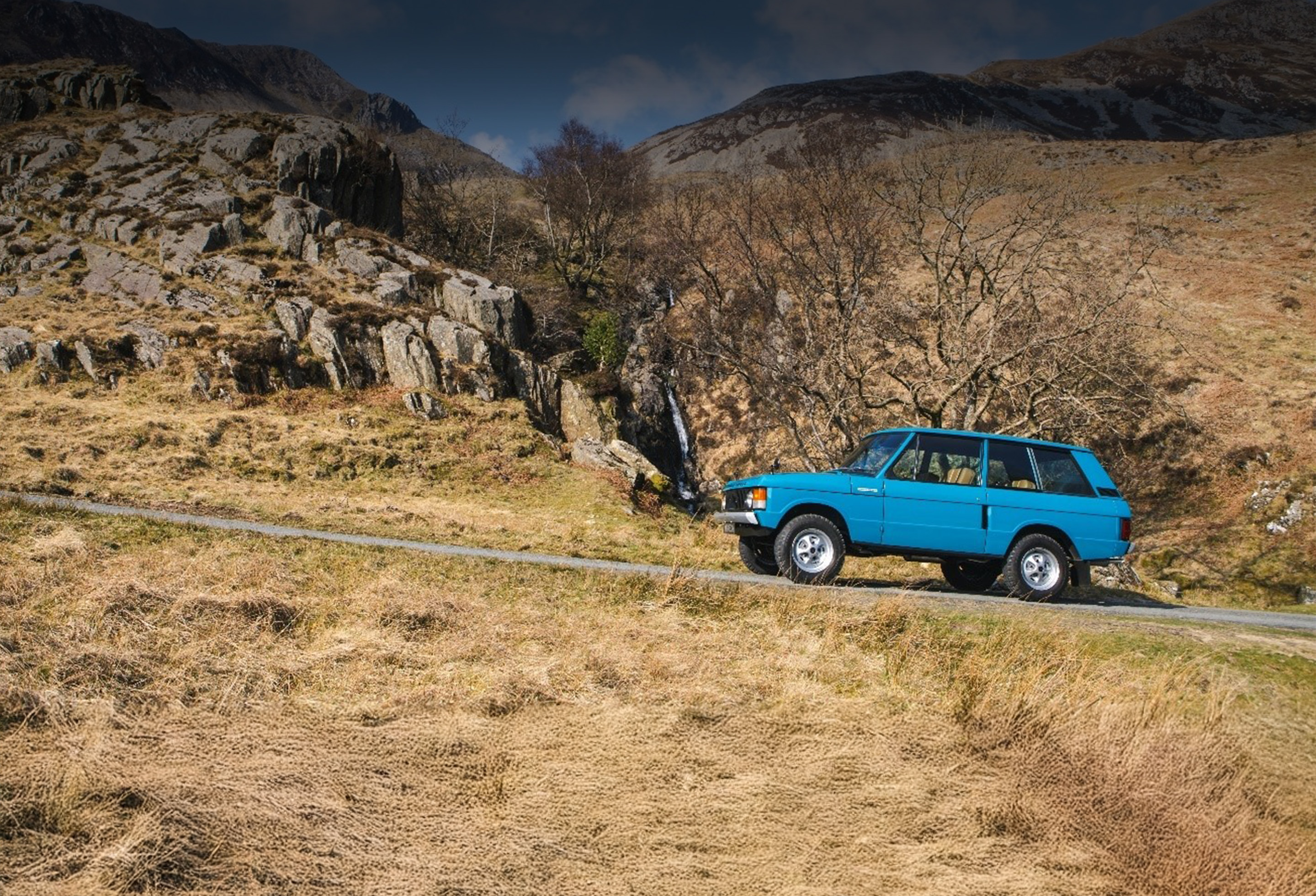 Blue Range Rover driving through rocky mountain road with waterfall in background.