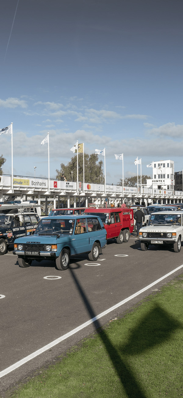 Lineup of vintage Land Rovers on racetrack with flags and viewing structure.